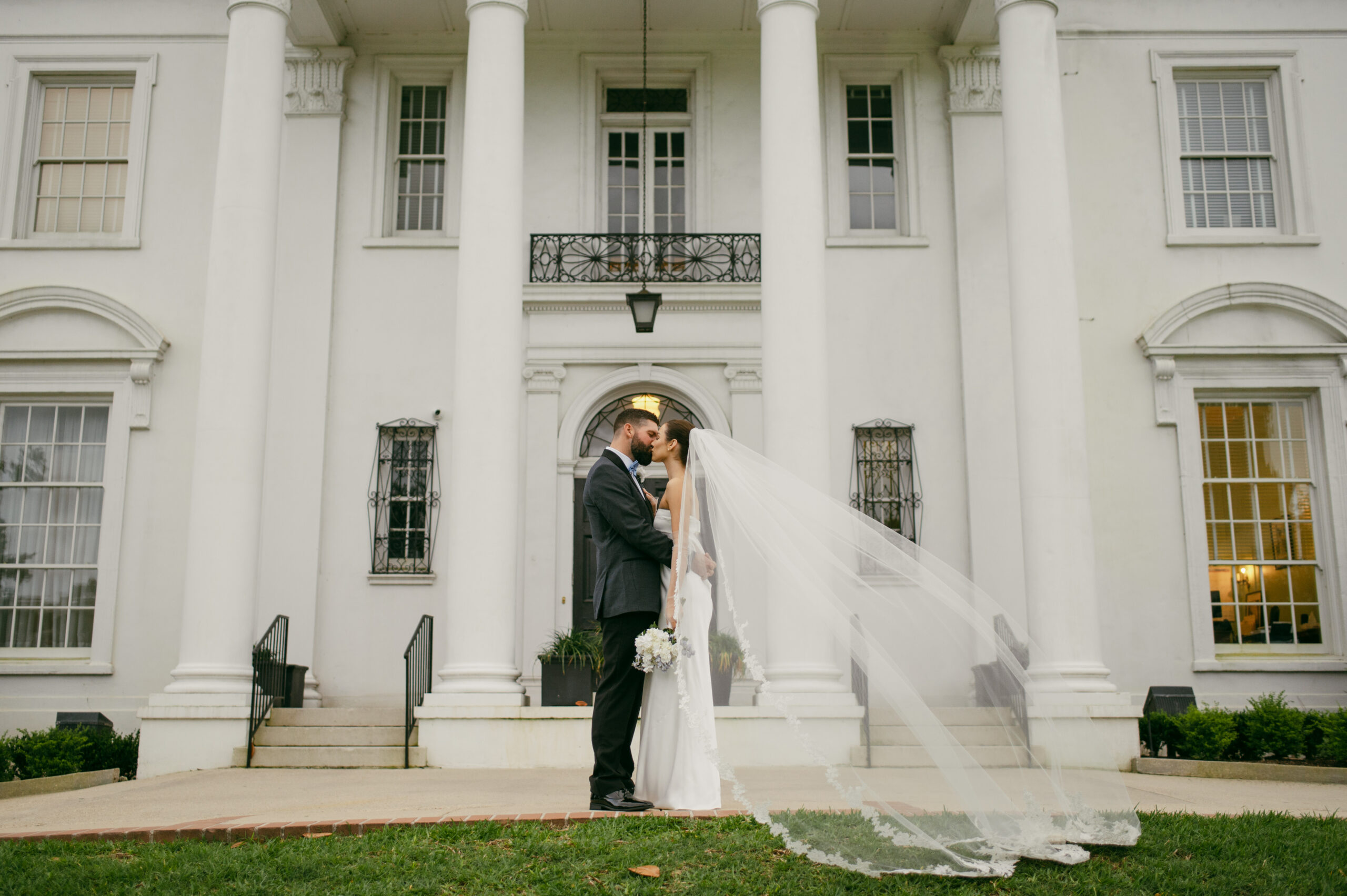 A bride and groom in front of the Old Governor's mansion as her veil blows in the wind.